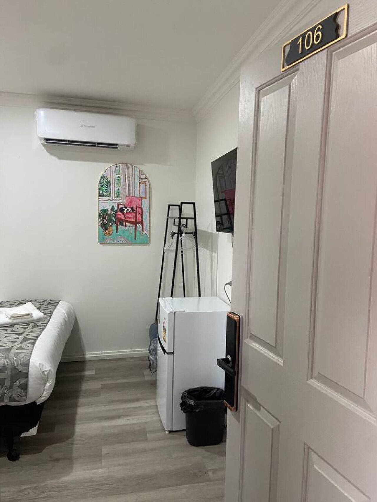 A room with a bed, a white refrigerator, a black trash bin, a painting on the wall, and a door with the number 106.
