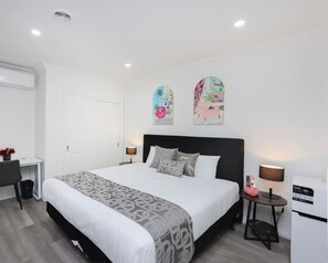 Ein modernes Hotelzimmer mit einem großen Bett, einem Schreibtisch und einem Stuhl. Ein kleiner Tisch mit einer Lampe und einem Mülleimer steht bereit. Die Wände sind weiß und die Decke verfügt über Einbauleuchten.