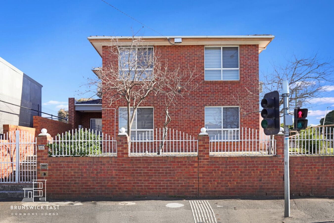 A two-story brick building with a white picket fence, a tree in front, and a traffic light.