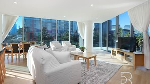Uma sala de estar moderna com um sofá grande, uma mesa de centro e uma TV. Há grandes janelas com vista para a cidade.