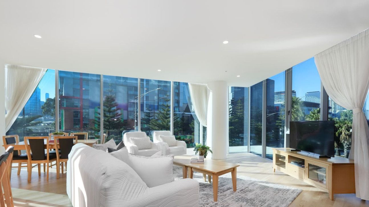 A modern living room with a large sofa, a coffee table, and a TV. There are large windows offering a view of the city.