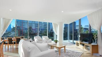 A modern living room with a large sofa, a coffee table, and a TV. There are large windows offering a view of the city.