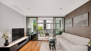 A modern living room with a flat-screen TV, a sofa, a dining table with chairs, and a balcony with plants. - Luxurious Docklands Escape| 1BR Apartment with Pool, Gym, Sauna, Balcony & Fast Wi-Fi (Docklands)