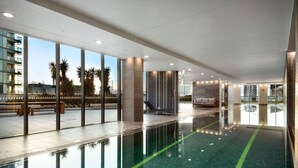 A modern indoor swimming pool with clear glass walls, a tiled floor, and a seating area with a sofa and a chair. - Luxurious Docklands Escape| 1BR Apartment with Pool, Gym, Sauna, Balcony & Fast Wi-Fi (Docklands)