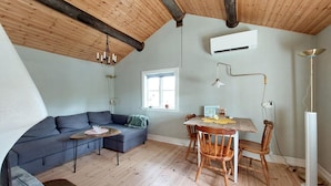 A cozy living room with a wooden ceiling, a gray sofa, a dining table with chairs, and a window providing natural light.