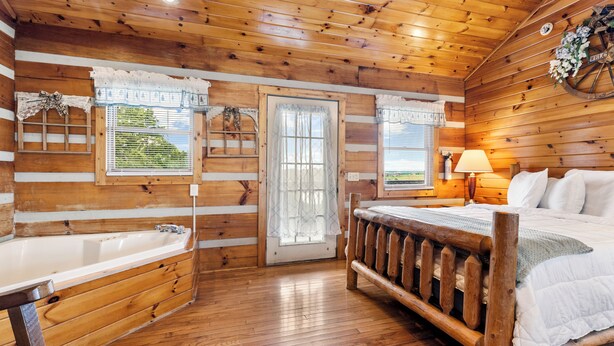 A wooden cabin bedroom with a large bed, a wooden bench, a bathtub, and a window with curtains. - Hillside Hideout Log Cabin For 2!  Cabin 10 (Fancy Gap)