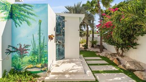 Une salle de bain extérieure moderne, dotée d’une douche à parois de verre, entourée de plantes tropicales et d’un jardin bien entretenu.