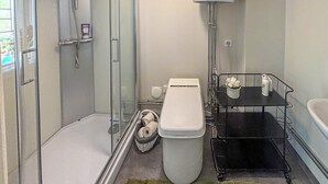 A modern bathroom with a glass shower enclosure, a toilet, and a black metal shelf unit with toiletries.