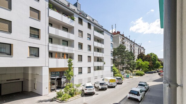 A multi-story residential building with balconies, a parking area, and a street view.