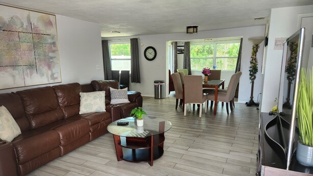 A living room with a brown leather sofa, a glass coffee table, a dining area with a table and chairs, and a large window with curtains.