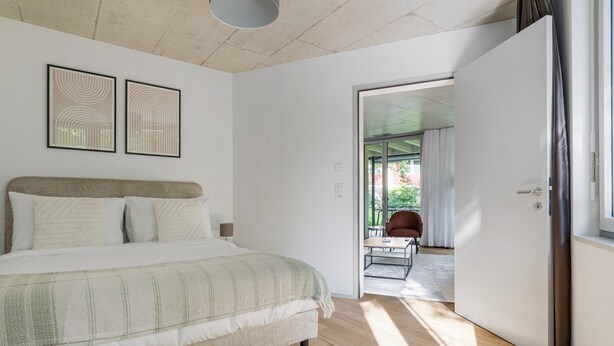 A bedroom with a bed, a chair, and a table. There is a door leading to another room. - Blueground | Saatlen, balcony, nr Zürich Zoo (Zurich)