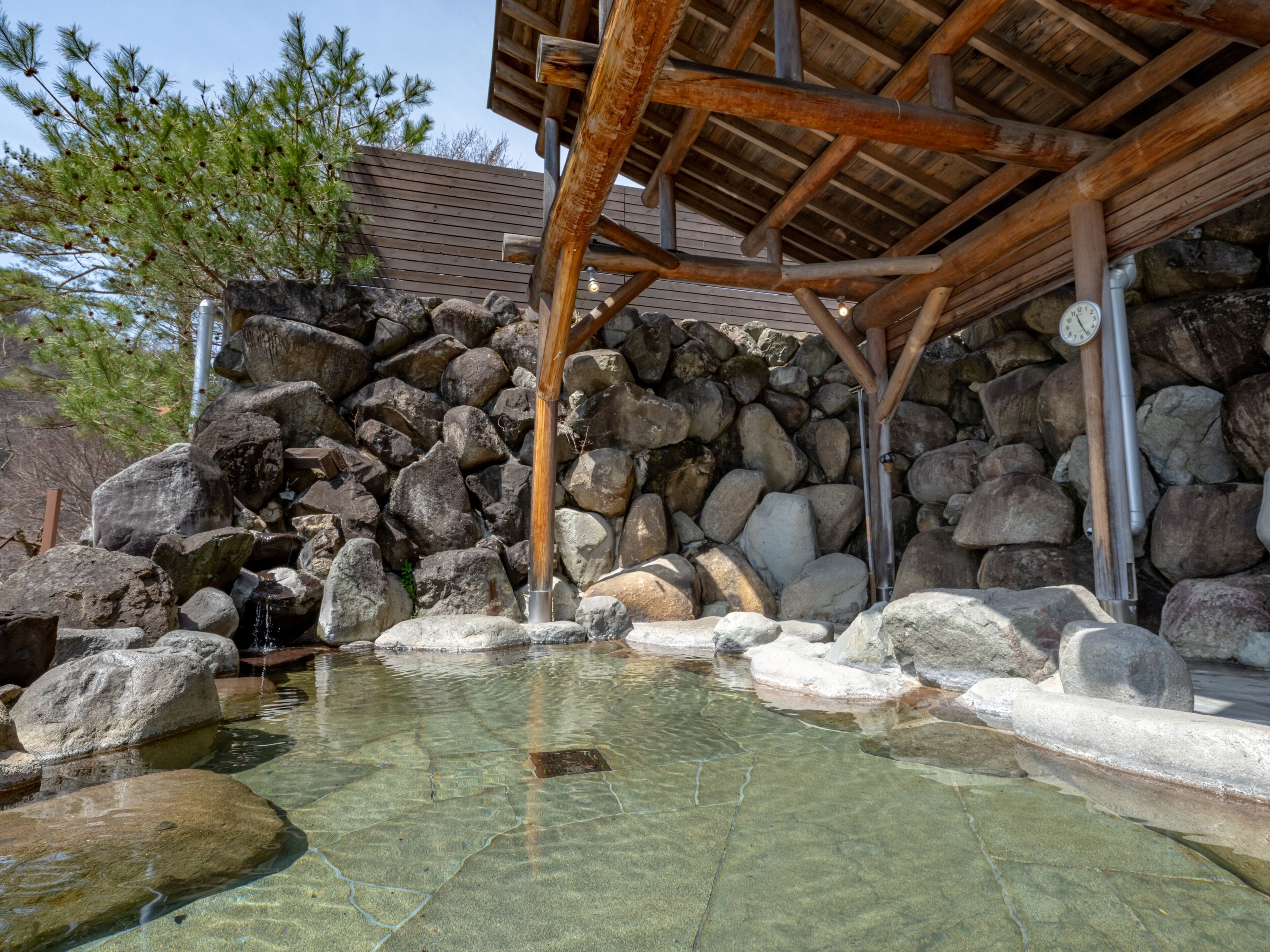 A traditional Japanese onsen with a wooden structure, a rock wall, and a clear hot spring.