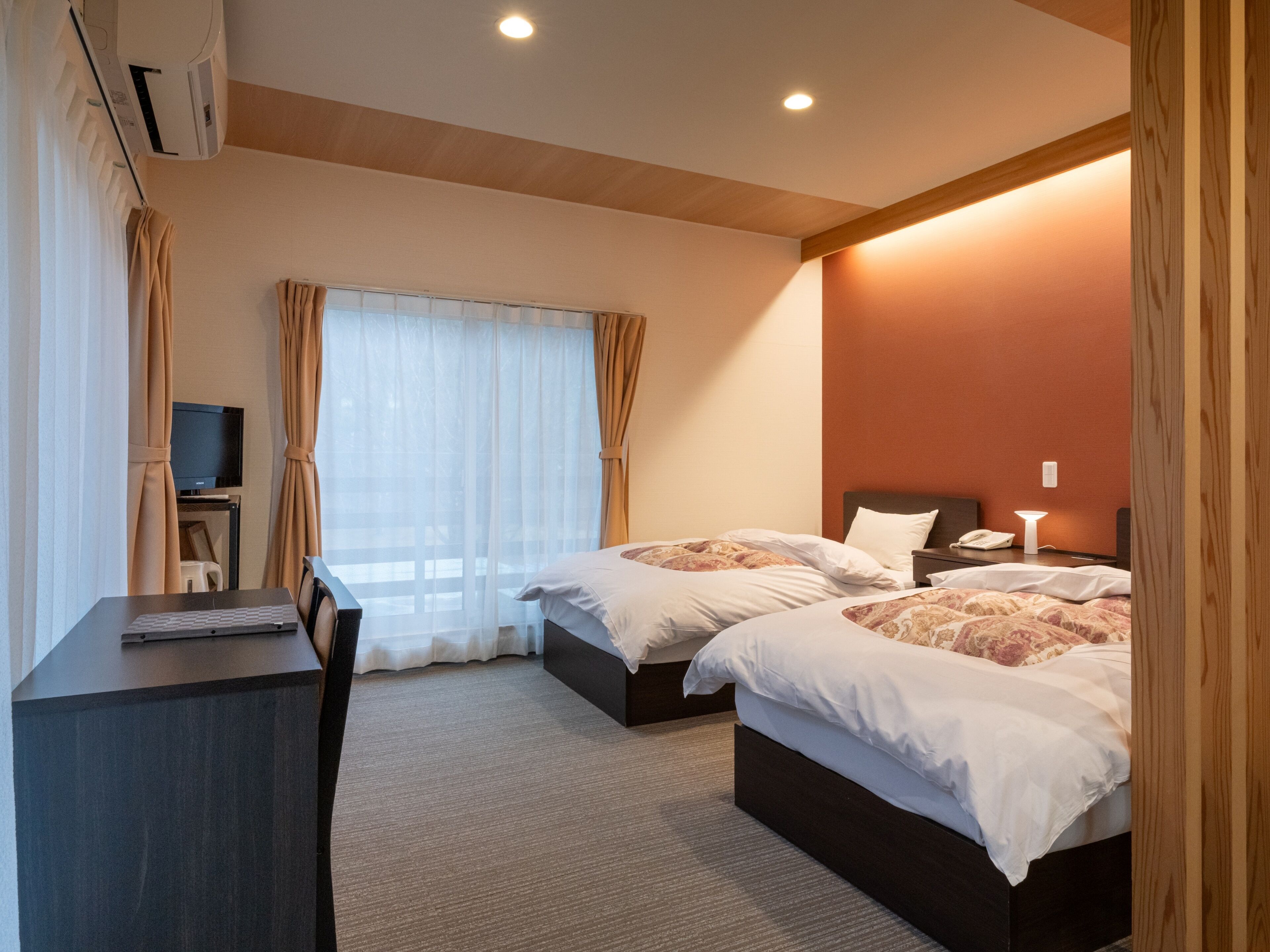 Una habitación de hotel con dos camas, un escritorio, televisión y ventana con cortinas.