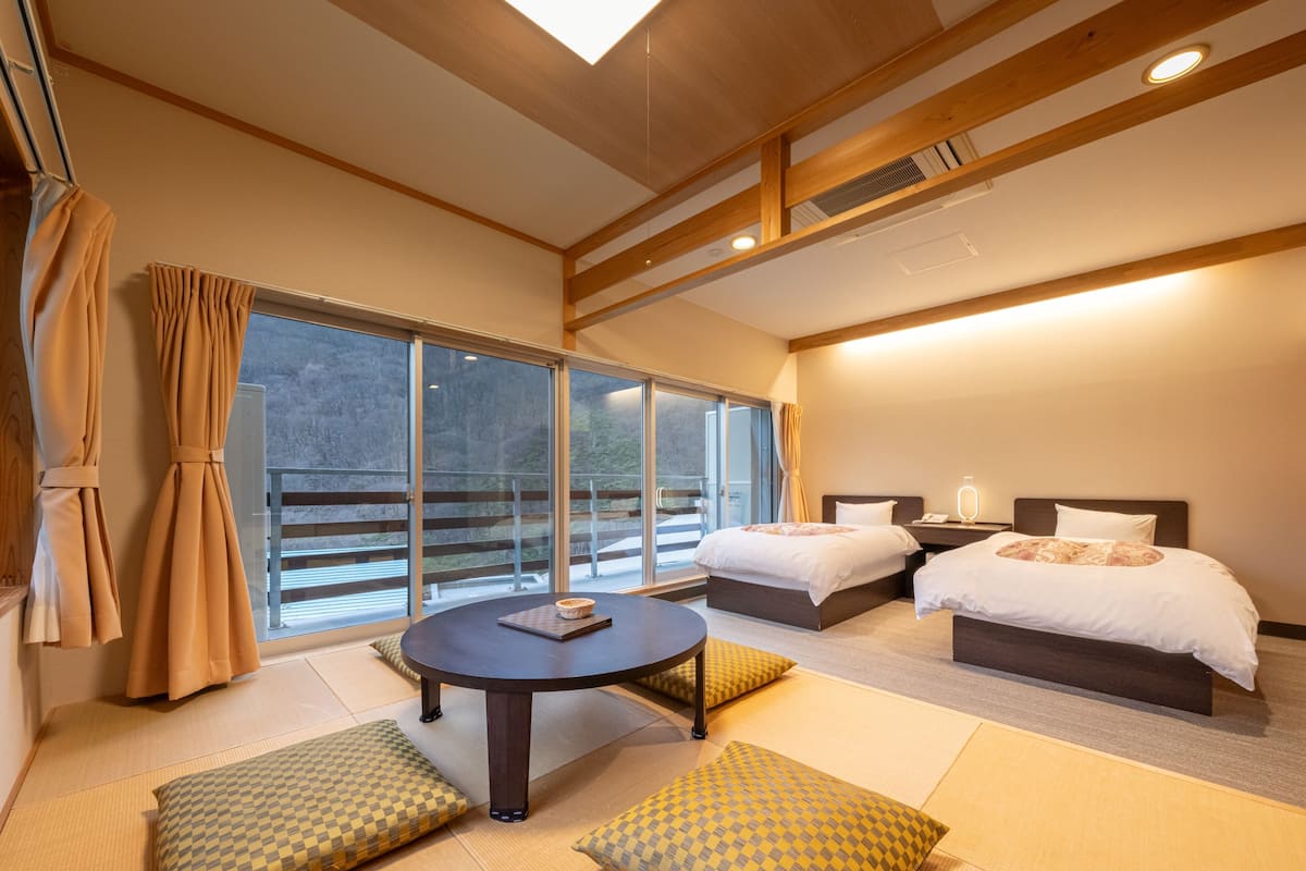 Una habitación de hotel moderna con dos camas, una mesa redonda y ventanas amplias que ofrecen vistas al exterior.