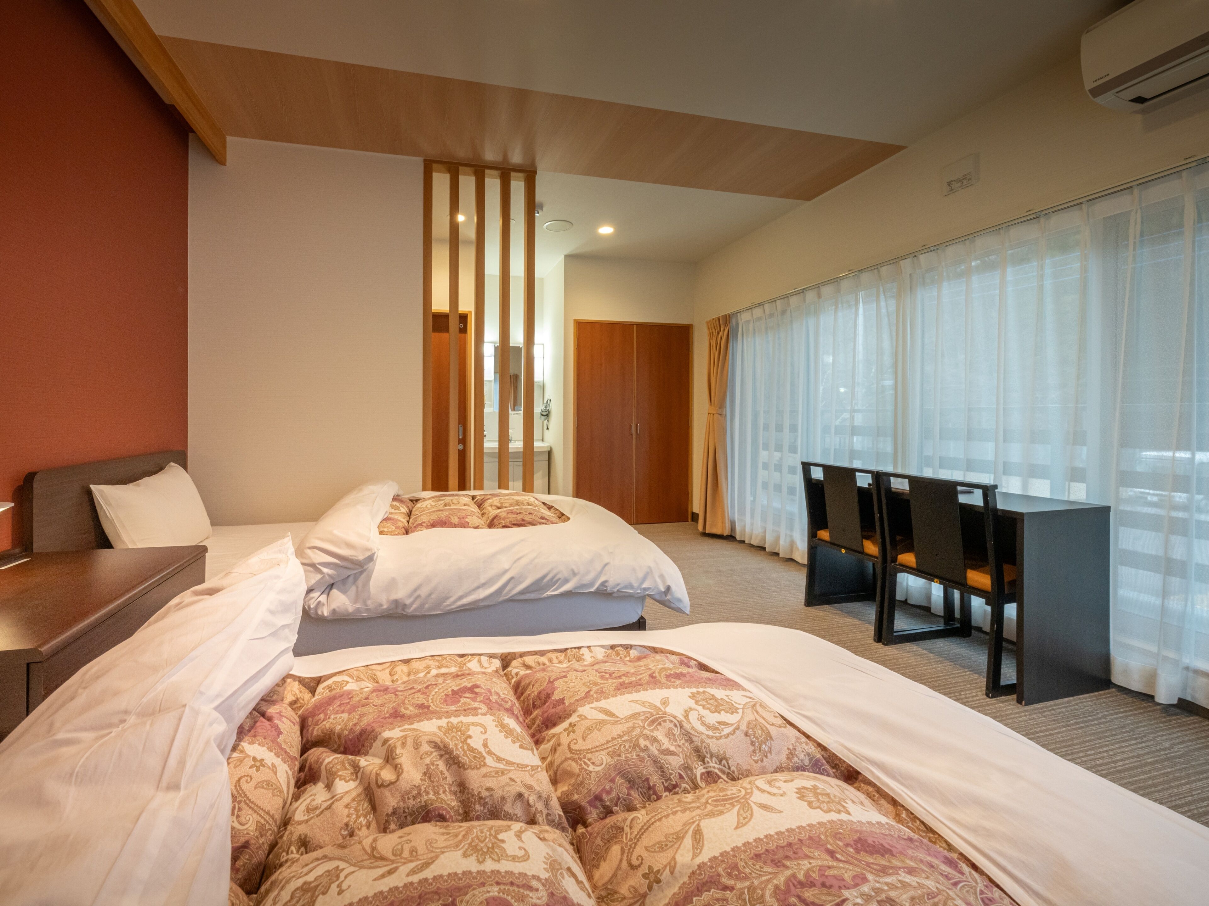 Une chambre d’hôtel avec un grand lit, un bureau et une chaise. Il y a une fenêtre avec des rideaux et un appareil de climatisation fixé au mur.