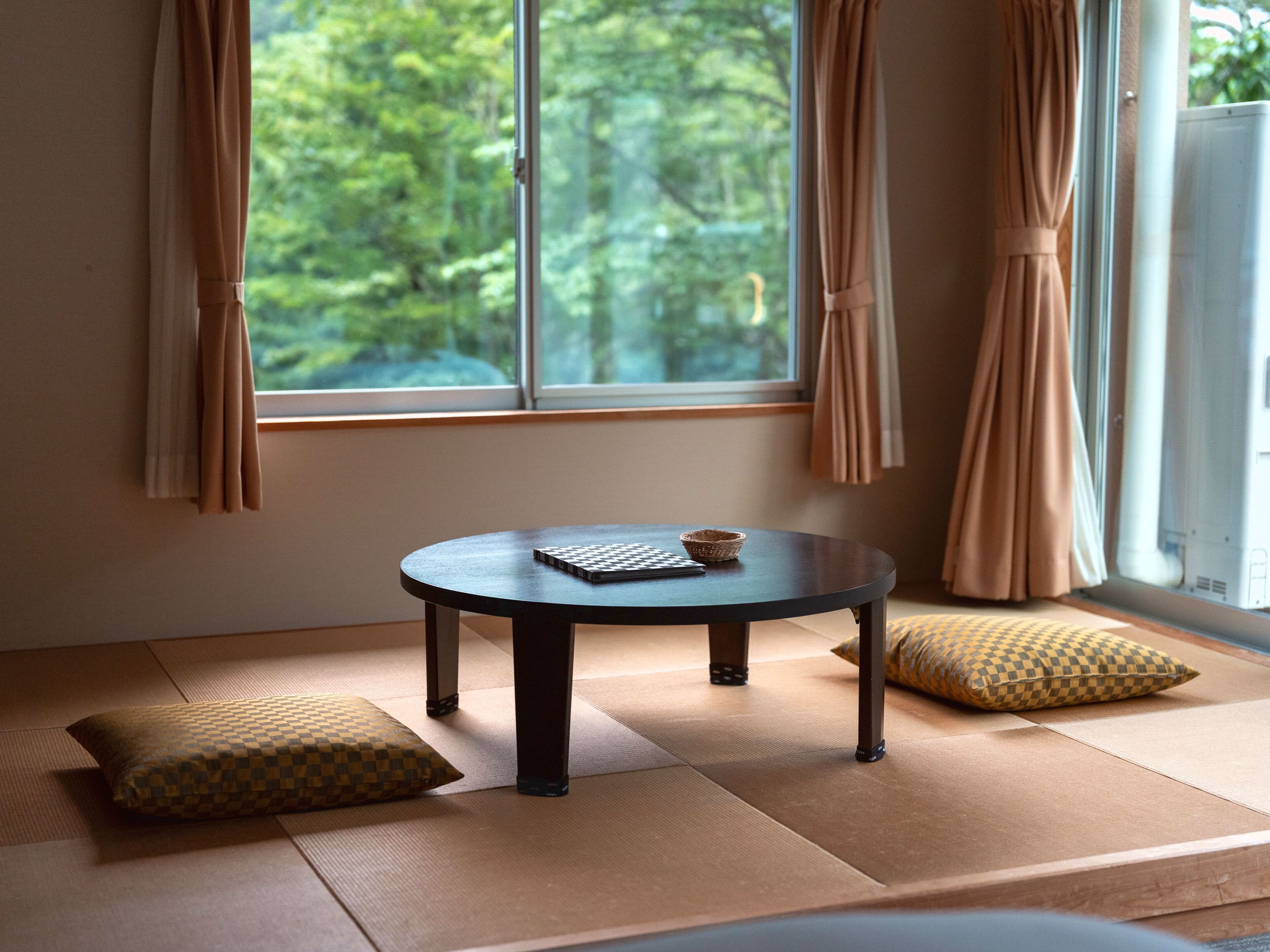 A tatami mat room with a round table, cushions, and a view of greenery through a window.
