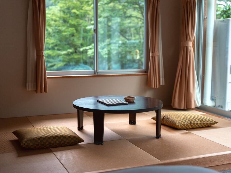 A tatami mat room with a round table, cushions, and a view of greenery through a window.