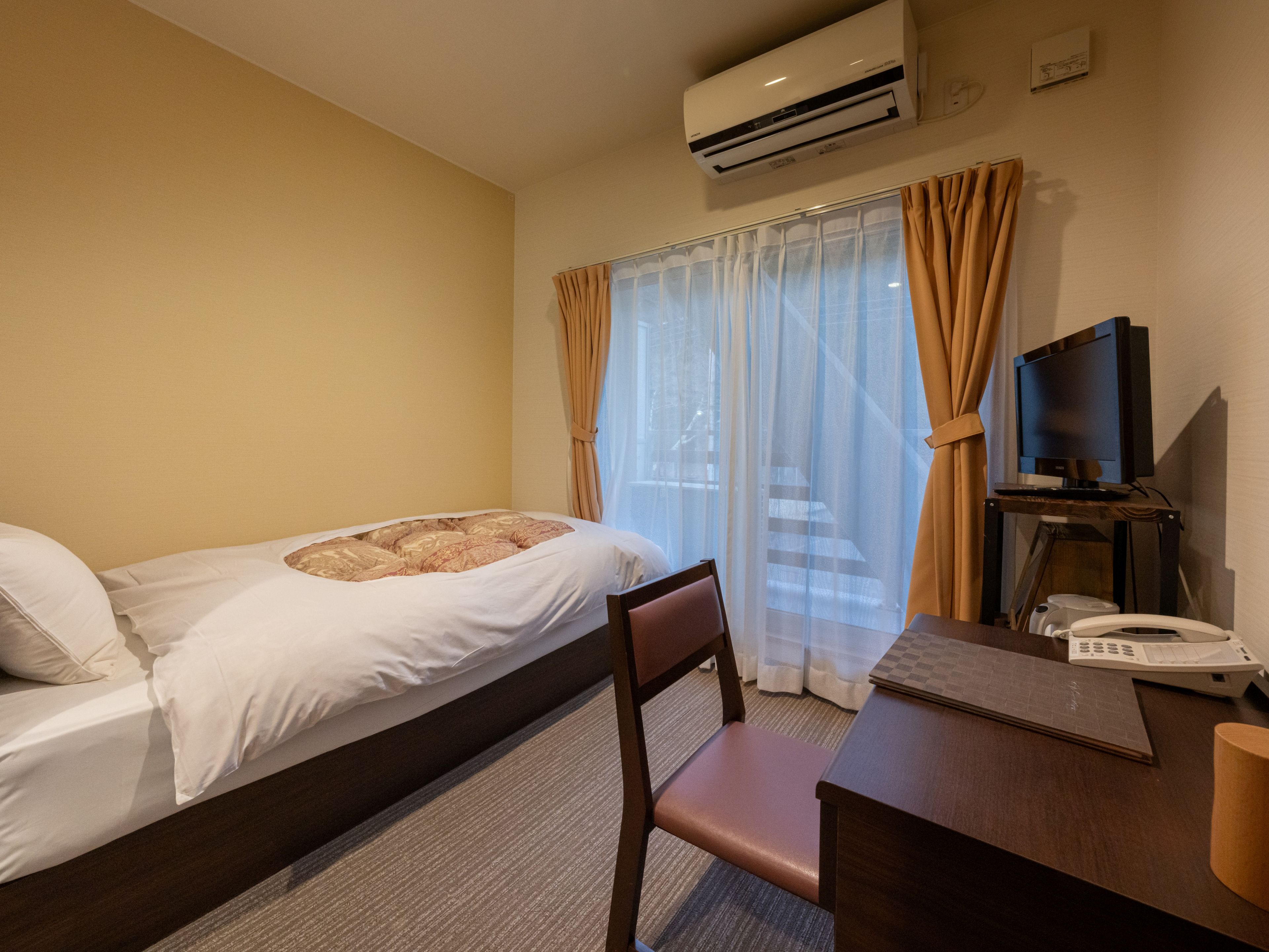 A hotel room with a bed, a desk, a chair, an air conditioner, a television, and a telephone.