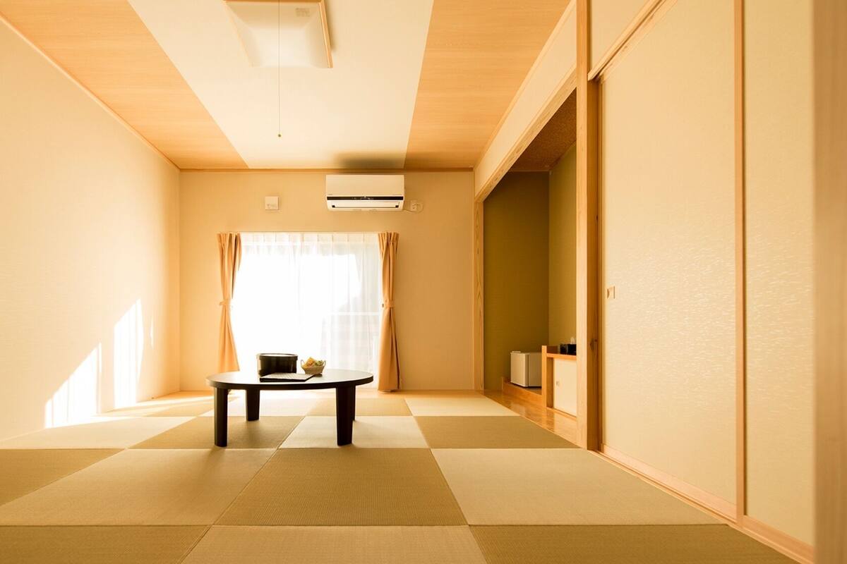 Una habitación con suelo de tatami, una mesita con un cuenco, un aire acondicionado blanco y una puerta corredera que da a otra habitación.