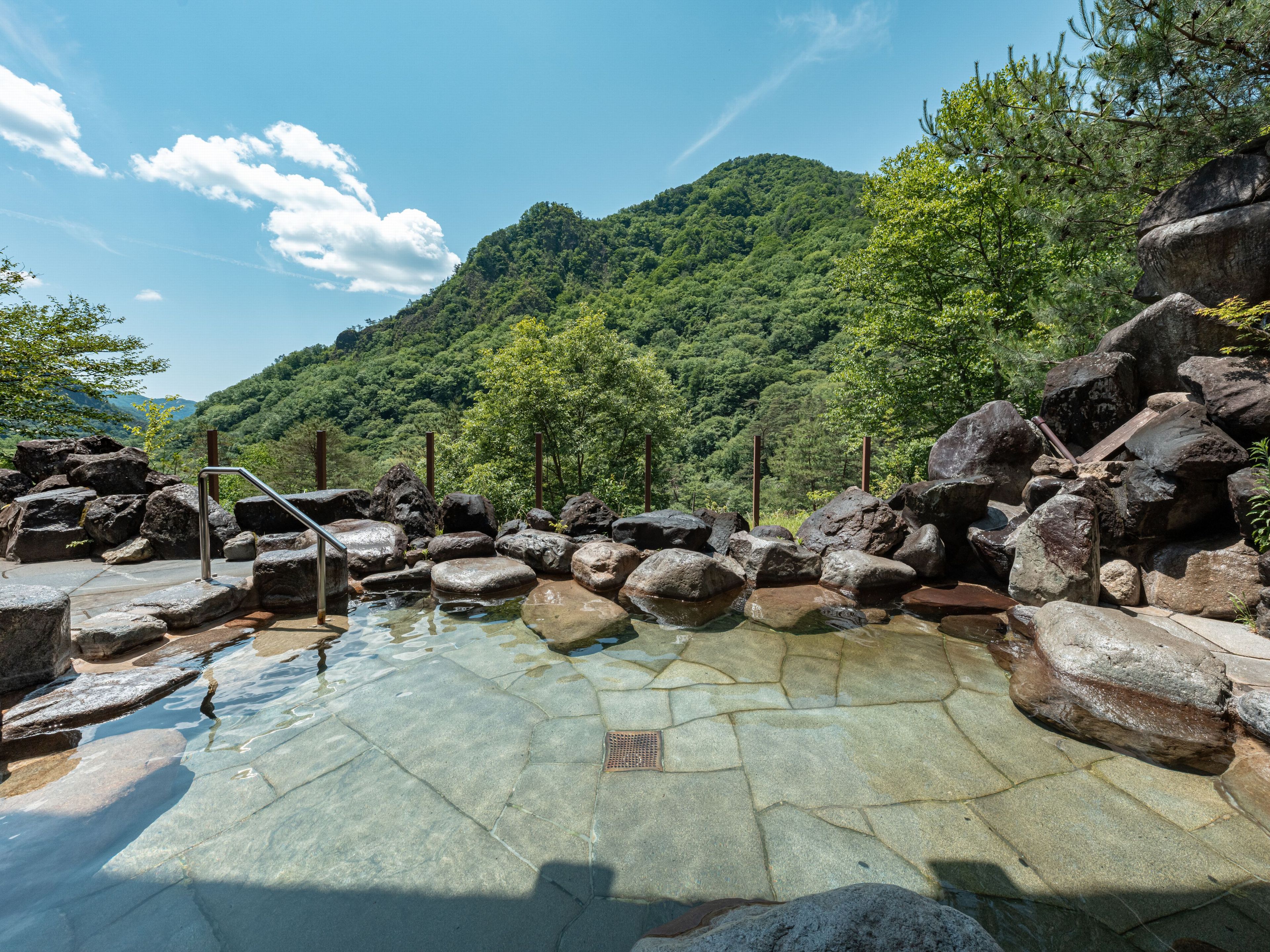 A natural hot spring surrounded by large rocks and trees, with a clear view of a mountain in the background.