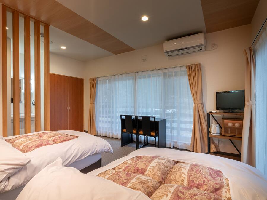 A hotel room with two beds, a desk with a television, an air conditioning unit, and a view through sheer curtains.