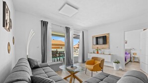 Una sala moderna con un sofá seccional gris, una mesa de centro de vidrio y un sillón amarillo. Se distingue un área de comedor visible a través de la puerta de vidrio, y un televisor montado en la pared.
