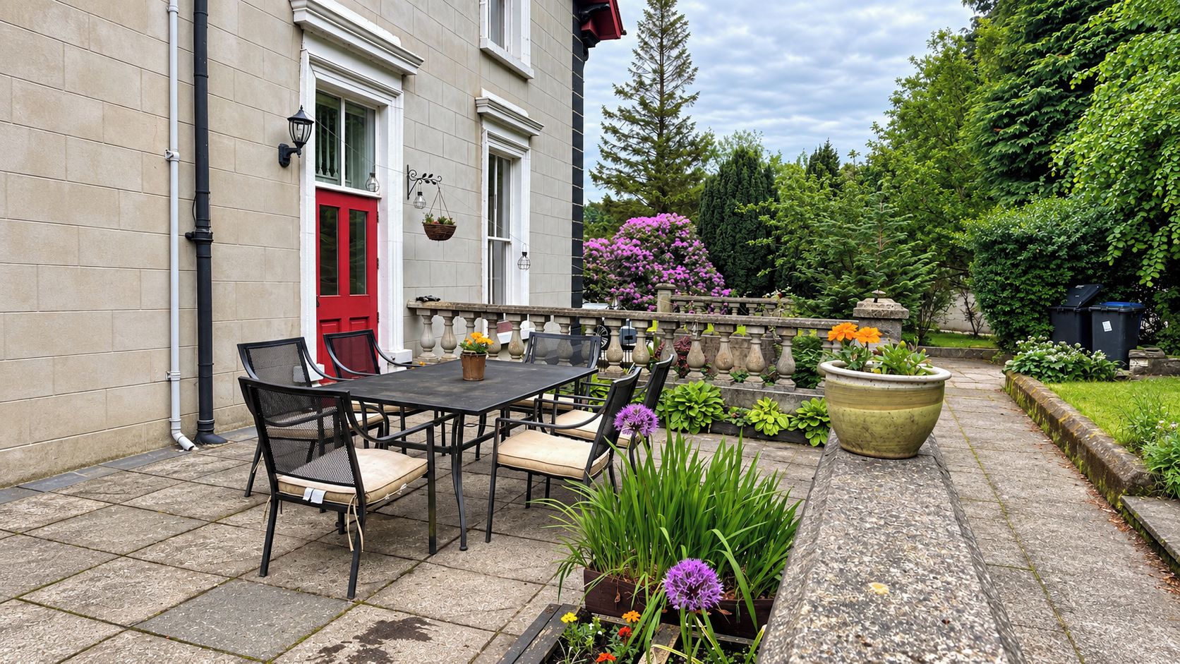 A patio with a table and chairs, a red door, and a garden with various plants and flowers.