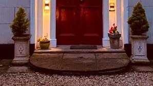 A building entrance with a dark red door, numbered 105, flanked by two potted plants and two wall-mounted lights.