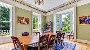 Comedor con una gran mesa de madera, sillas y una araña. Hay dos ventanas con vistas al follaje exterior.
