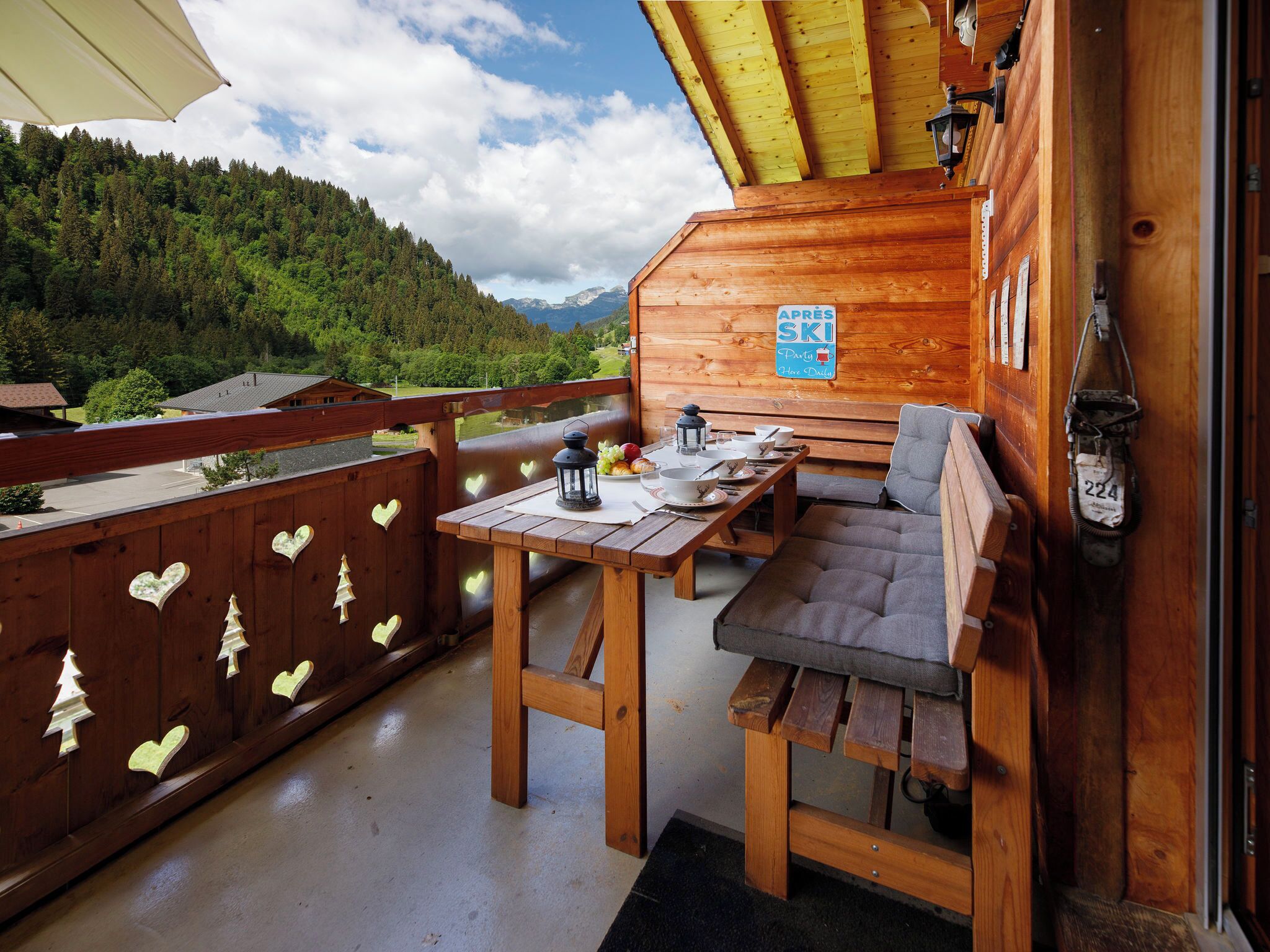 Eine Holzhütte mit Balkon, einem Tisch mit Stühlen, einem Schild mit der Aufschrift 'APRES SKI Party Hier Täglich' und Blick auf ein bewaldetes Gebirge.
