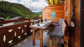 Eine Holzhütte mit Balkon, einem Tisch mit Stühlen, einem Schild mit der Aufschrift 'APRES SKI Party Hier Täglich' und Blick auf ein bewaldetes Gebirge.