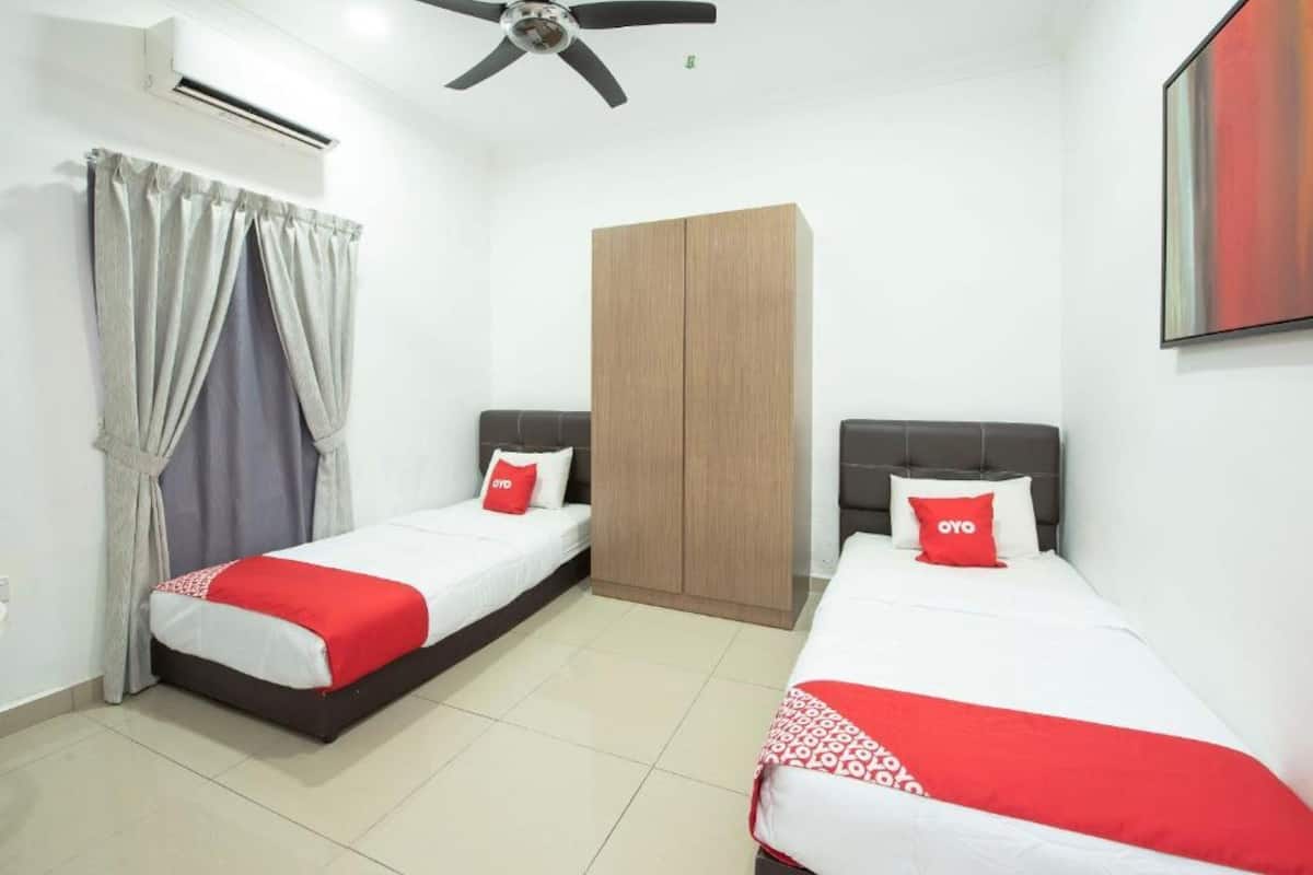 Habitación de hotel con dos camas individuales, cada una con cubrecama y almohadas rojas y blancas. Se observa un armario de madera, un ventilador de techo y un equipo de aire acondicionado.