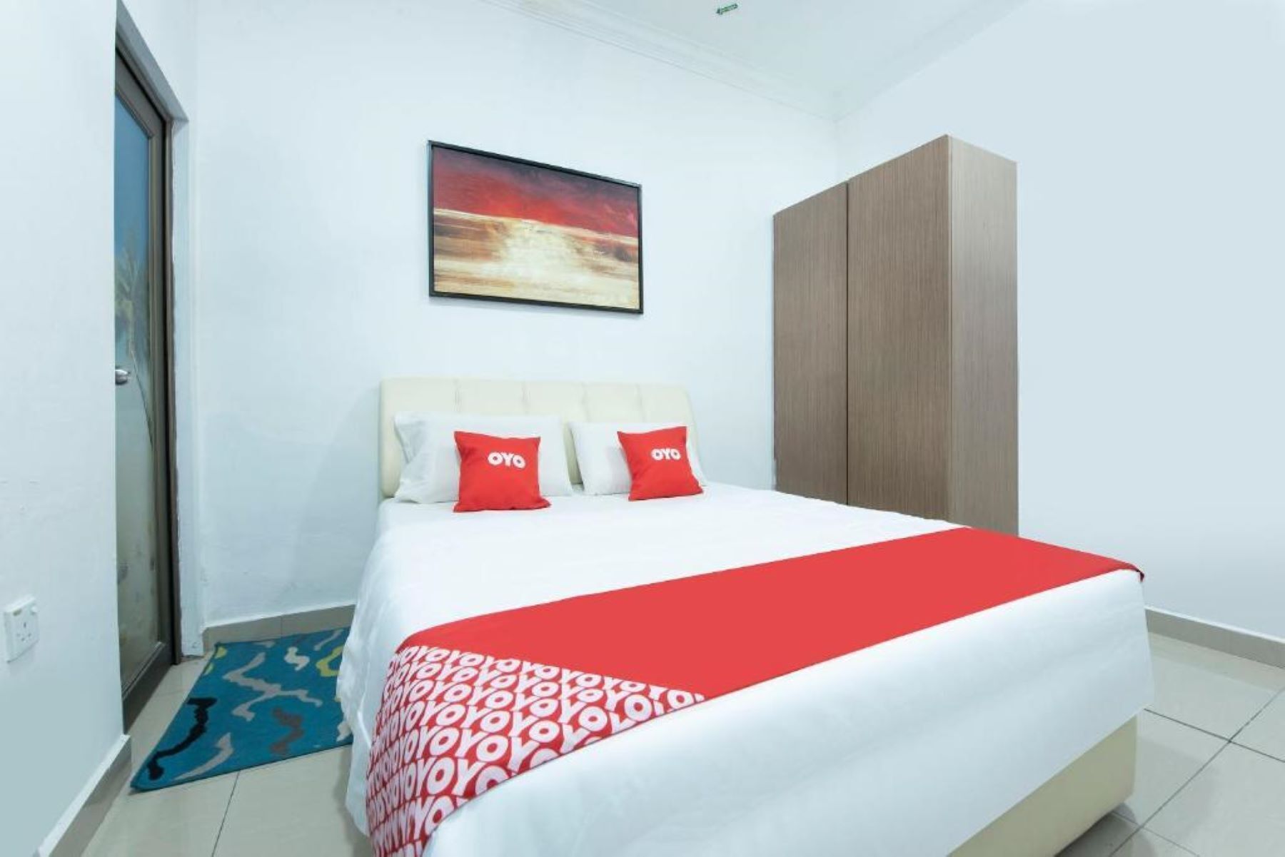 A hotel room with a bed, a red and white blanket, two red pillows with the logo, a painting on the wall, and a wardrobe.