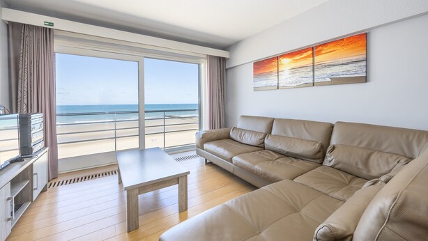 Un salon spacieux doté d’une grande fenêtre donnant sur une plage, d’un canapé en cuir beige, d’une petite table et d’un téléviseur à écran plat posé sur un meuble.