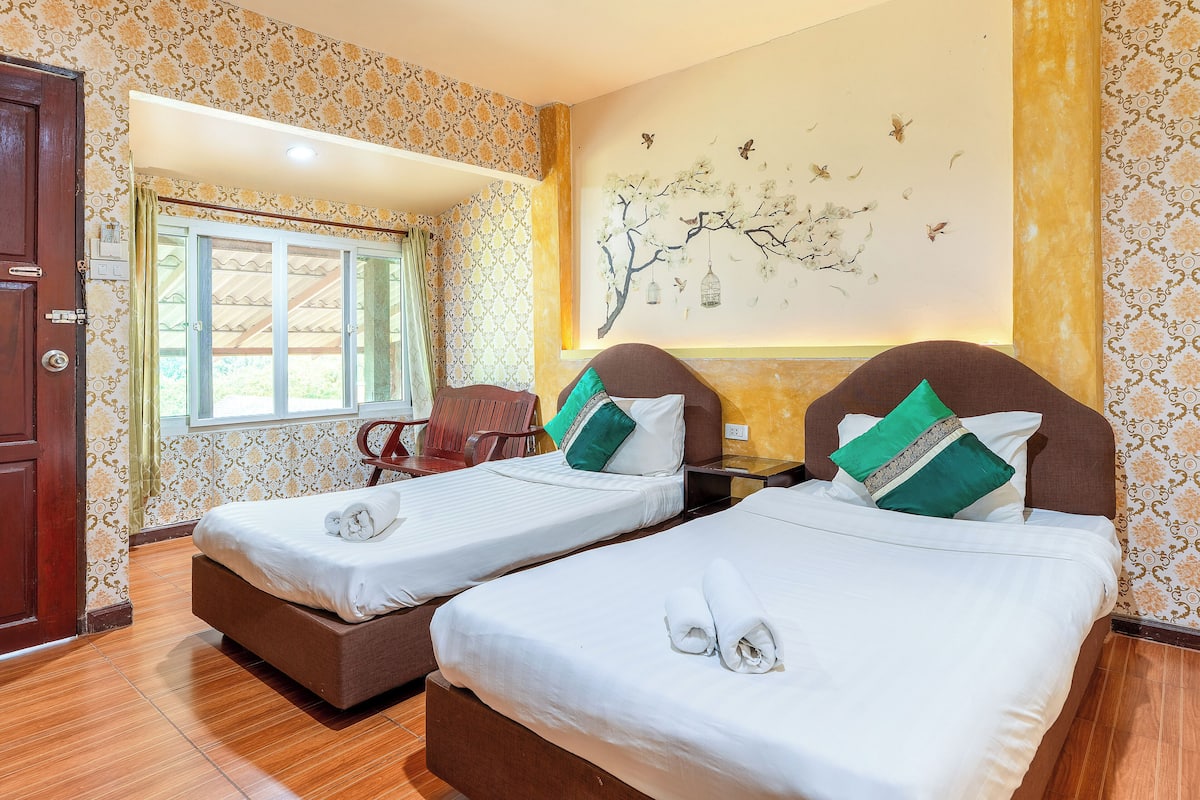 A hotel room with two single beds, a wooden door, a window with curtains, and a wall mural.