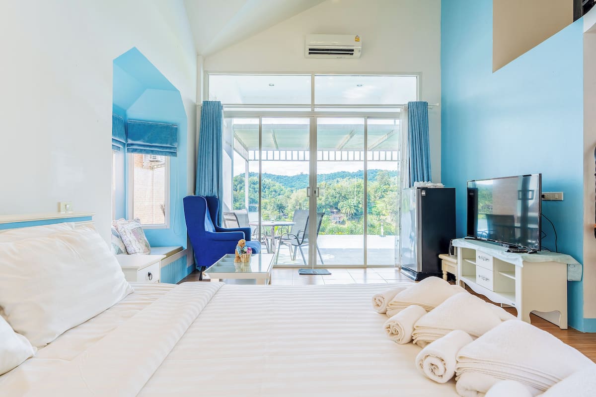 A hotel room with a large bed, a TV, a desk, and a balcony with a view of the greenery.