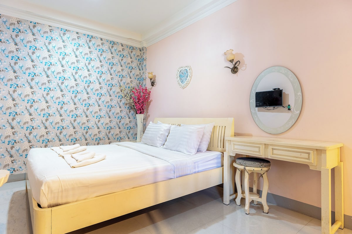 A neatly made bed with white linens, a small bedside table with a chair, a mirror, and a wall with floral wallpaper.