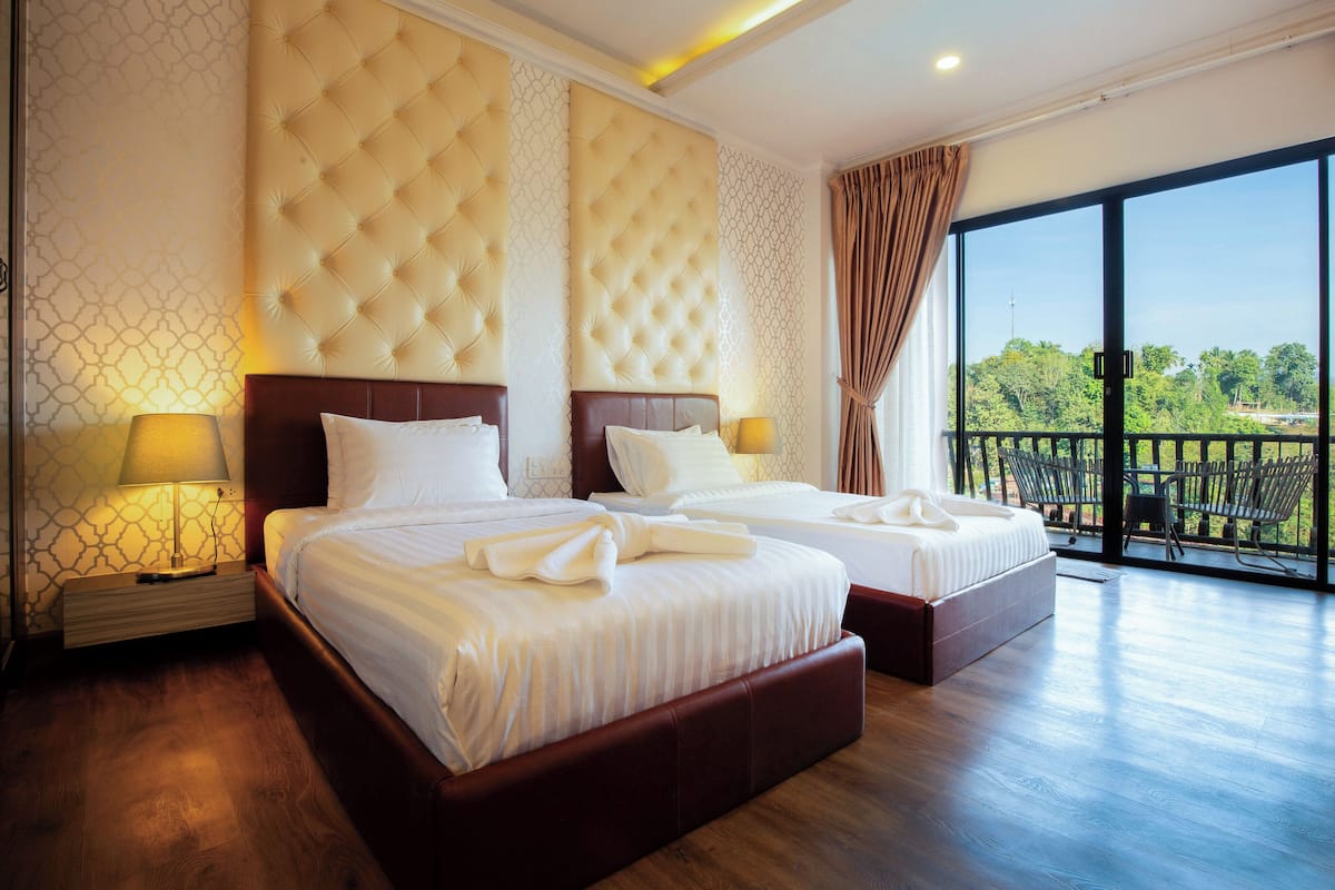 A hotel room with two beds, a large window with a view, and a balcony with outdoor furniture.