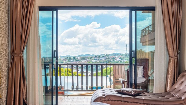 A bedroom with a large bed, a view of a balcony with a pool, and a cityscape in the background. - Samprasob Resort (Sangkhla Buri)