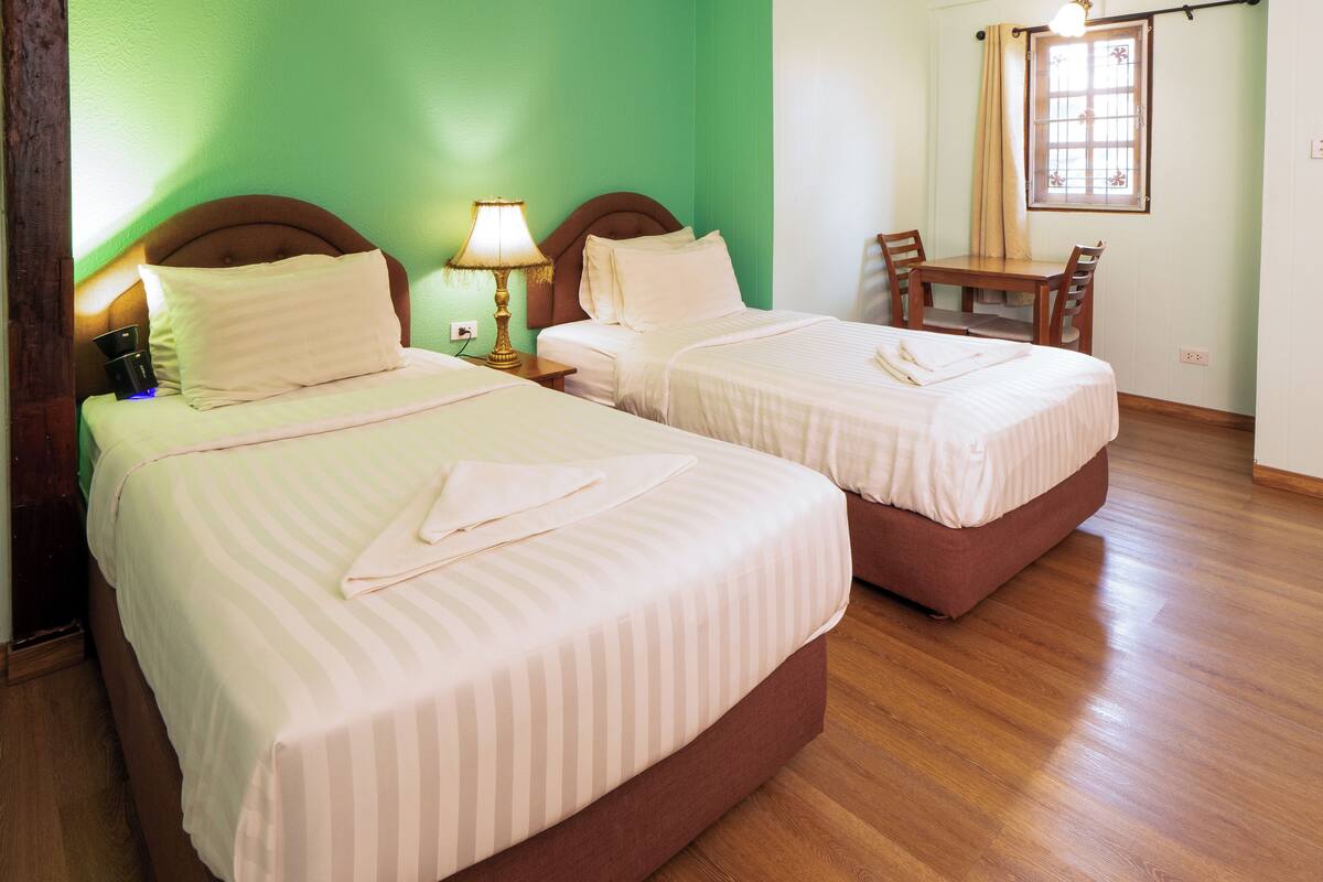 Two beds with striped bedding, a bedside lamp, a wooden table with chairs, and a window with curtains.
