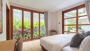 A hotel room with a bed, a chair, a desk, and large windows offering a view of a garden.