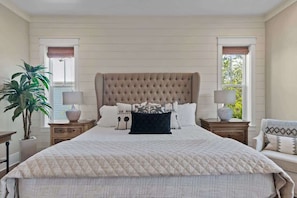 Quarto com cama grande, duas mesas de cabeceira, um criado-mudo, uma planta em vaso e janela com vista.
