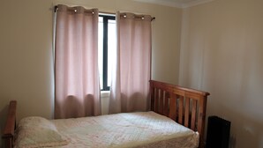 A single bed with a wooden headboard in a room with beige walls and a window with curtains.