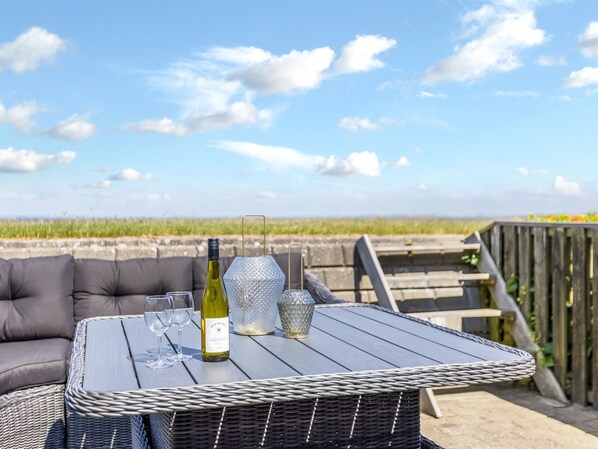 A wicker table with a bottle of wine and glasses, set on an outdoor deck with a view of the beach.