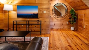 Interior acolhedor de uma cabana de madeira, com televisão, luminária, espelho e planta em vaso.