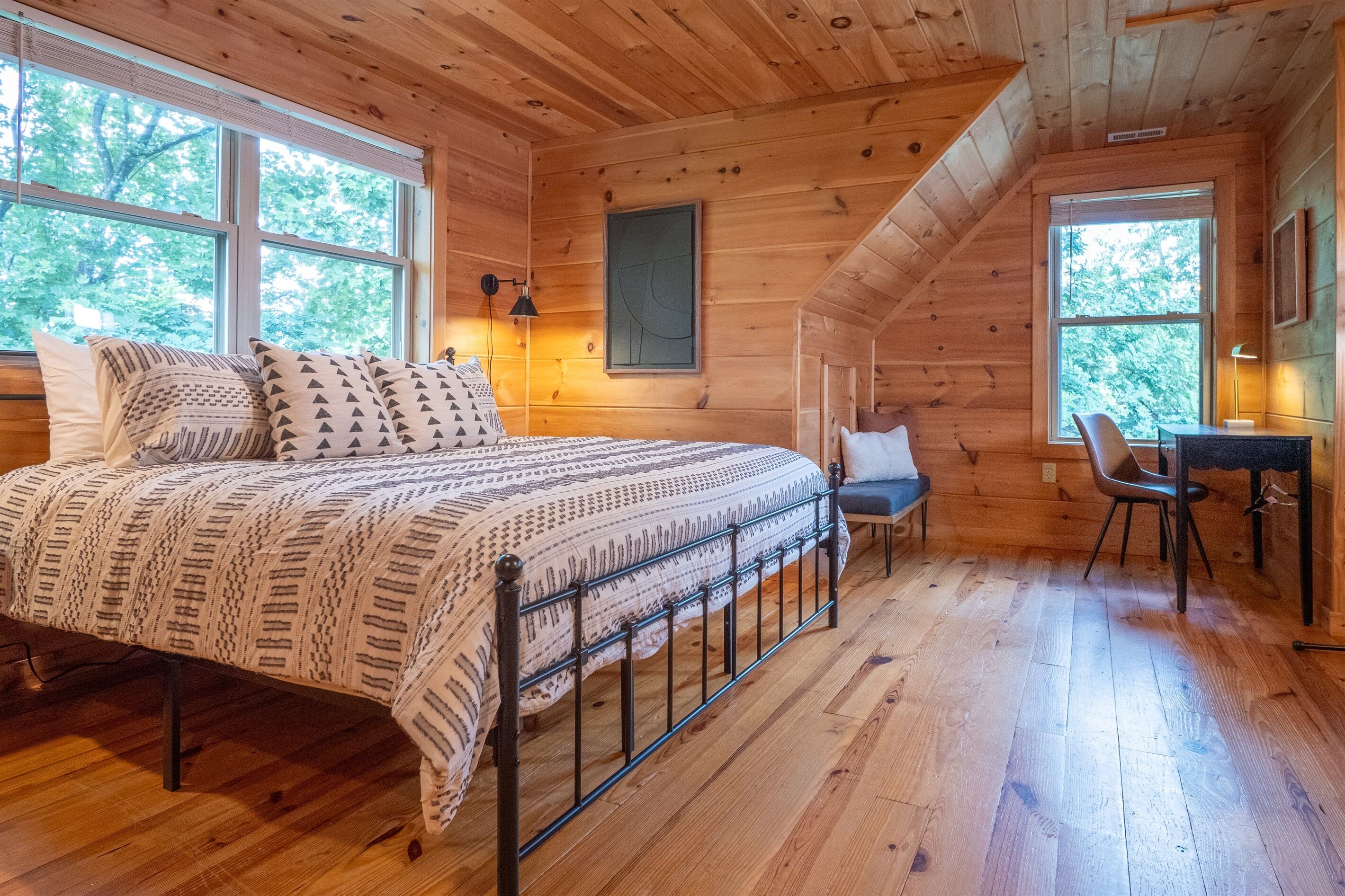 Dormitorio de una cabaña de madera con una cama, dos ventanas, un escritorio y una silla.