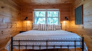 Un dormitorio de cabaña de madera con una cama, dos lámparas de pared y una ventana con vistas a árboles.