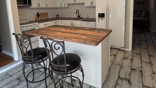 Een keuken met witte kastjes, een houten kookeiland met twee stoelen, een roestvrijstalen koelkast en een plafondlamp.