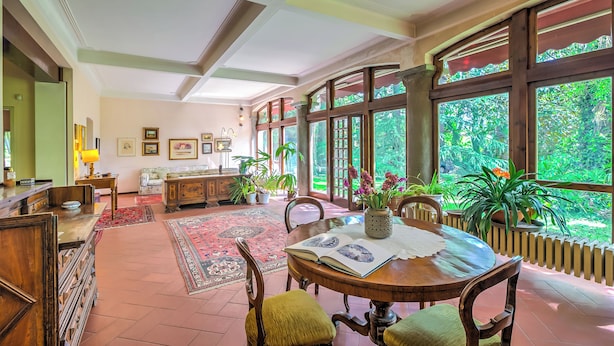 A spacious room with a large wooden dining table, several chairs, and a view of lush greenery through large windows.