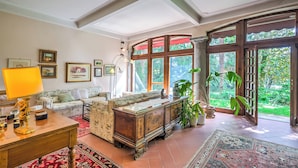 A spacious living room with a large wooden cabinet, a sofa, and a coffee table. There are several framed pictures on the wall and a large window providing a view of the outdoors.